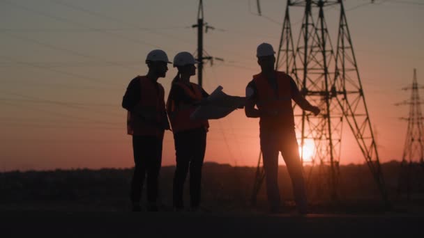 Utveckling av elektrisk struktur i staden, professionella ingenjörer i hjälmar diskuterar byggandet av elektriska torn bakgrund av solnedgång, siluett — Stockvideo