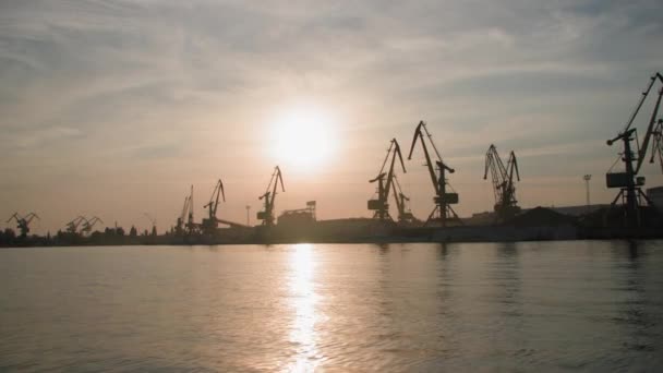 Силует, міський морський порт з підйомними кранами для завантаження вантажів на морські торговельні судна на тлі заходу сонця, вид з річки — стокове відео