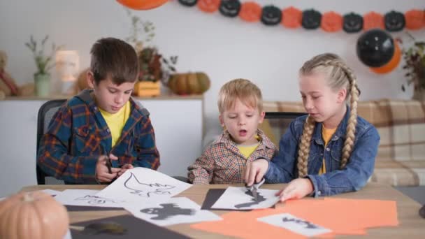 Halloween thuis, kinderen met een schaar gesneden figuren van geest en vleermuis zitten aan tafel tegen de achtergrond van decoraties en ballonnen — Stockvideo