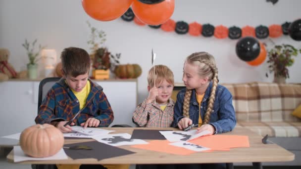 Préparant des décorations pour la fête d'Halloween à la maison, les enfants avec des ciseaux découpent des figures de fantôme et de chauve-souris sur des guirlandes et des ballons de citrouille de boulangerie — Video