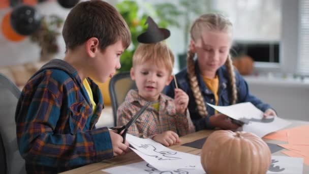 Хлопчик і дівчинка з ножицями вирізають статуетки привида і кажана для Хеллоуїна, маленька дитина тримає капелюх на паличці в руках, сидячи за столом з гарбузом — стокове відео