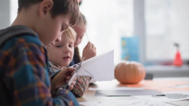 Közelről gyermek ollóval vágott egy fekete denevér papírból a háttérben a gyerekek ül az asztalnál tök, házi készítésű dekorációk a Halloween ünnep — Stock videók
