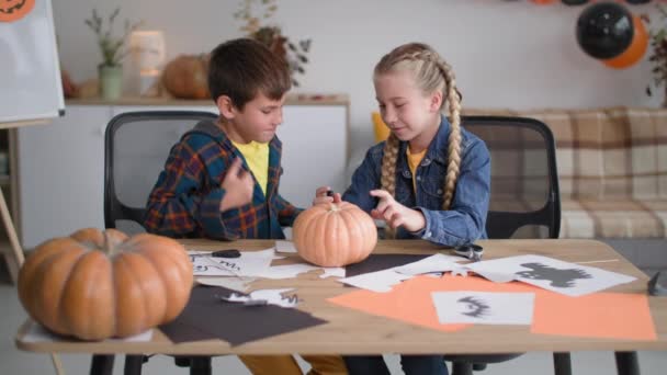 Przygotowanie do Halloween, dzieci z końcówką filcu narysować twarz na dyni i pokazać kciuki do siebie na tle girlandy i balonów — Wideo stockowe