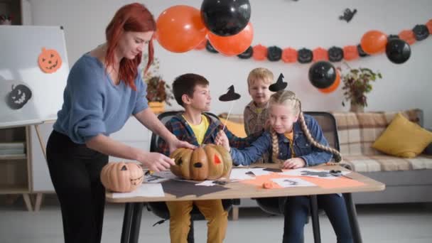 Decoraciones de Halloween caseras, mamá ayuda a los niños a cortar la calabaza, familia sentada en la mesa se divierten juntos en el fondo de guirnaldas y globos — Vídeos de Stock