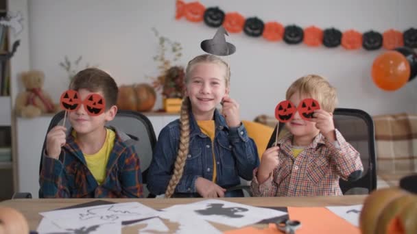 Niños felices celebran halloween, niños con máscaras en forma de calabazas y niña con un sombrero en un palo dice boo y se ríe en casa — Vídeo de stock