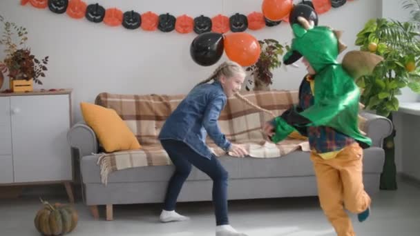 Chłopiec w smoczym kostiumie biegnie za dziewczyną po pokoju ozdobionym pomarańczowymi i czarnymi balonami i girlandami w kształcie dyń — Wideo stockowe