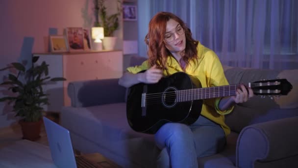 Vrouwelijke instructeur leert online met behulp van laptop om gitaar te spelen terwijl thuis zitten in de avond — Stockvideo