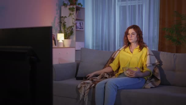 Домохозяйка любит смотреть телевизор и есть попкорн, сидя на диване, молодая женщина эмоционально реагирует на кино во время просмотра во время отдыха в гостиной — стоковое видео