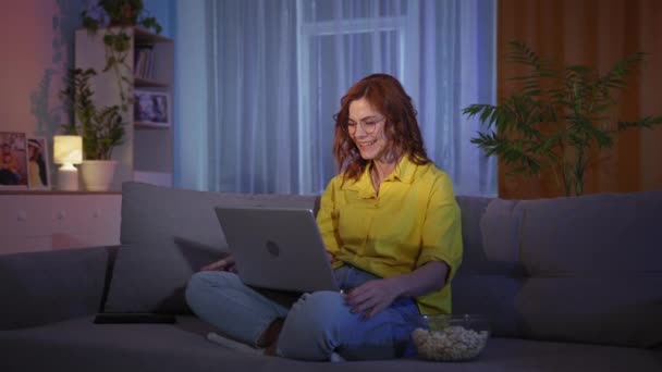 Емоційна щаслива дівчина радіє виграшем або хорошими новинами в інтернеті на дивані з ноутбуком і попкорн вдома — стокове відео