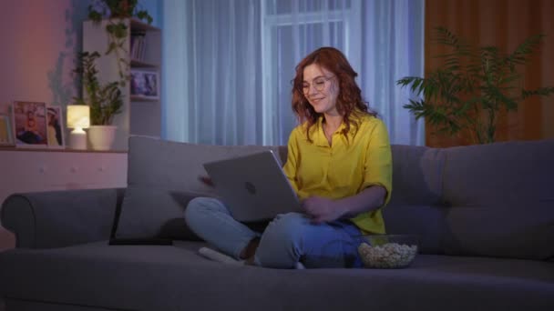 Woman eats popcorn and joyfully reacts to good news or won on Internet while sitting on couch with laptop on couch in evening — Vídeo de Stock