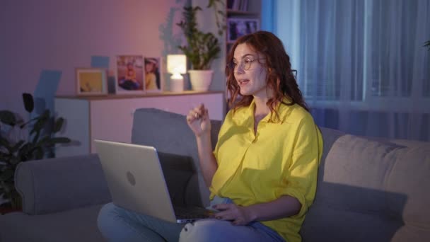 Kobieta wykorzystuje nowoczesne technologie i rozmowy przez kamerę internetową na laptopie podczas siedzenia na kanapie w salonie wieczorem, pobyt w domu — Wideo stockowe
