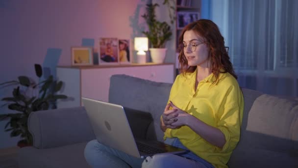 Vrouw met bril gesprekken met vrienden online met behulp van videocamera op laptop en internet, terwijl zitten op de bank thuis in de avond tijdens sociale afstand — Stockvideo