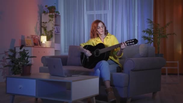 眼鏡をかけた若い女性はリビングルームで夜ソファに座っているラップトップでオンラインギターのレッスンを受けます — ストック動画