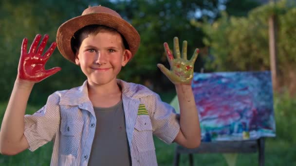 Kinderen entertainment, vrolijke mannelijke kind toont zijn handpalmen in verf om camera terwijl het tekenen op doek in de open lucht in het park achtergrond van groene bomen — Stockvideo