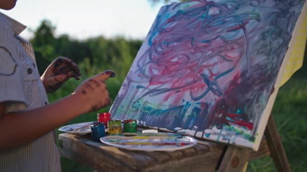 Küçük erkek sanatçı parmaklarıyla boyayı alıyor ve tuvale bir resim çiziyor, dışarıda dinlenirken, yakın plan. — Stok video