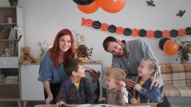 Fiesta de otoño padres felices con su diversión haciendo decoración de papel para Halloween y gafas de burbuja calabaza, sonriendo y olas a la cámara mientras están sentados en la mesa en la habitación festiva — Vídeo de stock