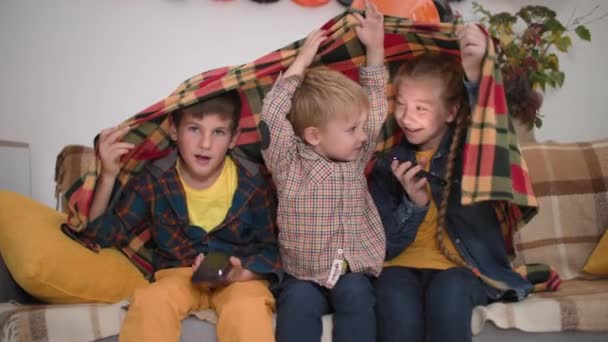 Halloween parties, funny kids with phones in their hands shine on their faces and tell scary stories covered with blanket while sitting on couch in party room — Vídeo de Stock