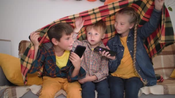 Leuk tijdverdrijf voor kinderen, vrolijke jongens en meisje met telefoons in hun handen schijnen op hun gezichten en vertellen enge verhalen bedekt met deken lach en kijk naar de camera, terwijl zitten op de bank in partij — Stockvideo