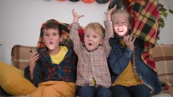 Παιδιά Απόκριες παιχνίδια, πορτρέτο των αξιολάτρευτο αγόρια και κορίτσια με μακριά ξανθά μαλλιά κάθεται στον καναπέ κάτω από κουβέρτα και να τους τρομάξει ουρλιάζοντας boo, χαμογελώντας και κοιτάζοντας κάμερα — Αρχείο Βίντεο