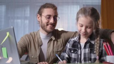 Baba, uzaktan öğrenme sırasında video bağlantısı yoluyla öğretmenle ödev yapmasına yardım ediyor, kameraya bakarken eğleniyor ve dans ediyor.