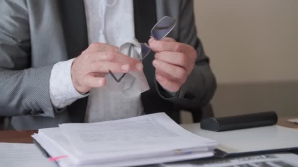 Человек в костюме вытирает очки салфеткой во время чтения документов в офисе, крупным планом — стоковое видео