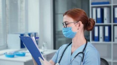 Mavi üniformalı güzel bir kadın doktorun portresi steteskopla not yazıyor. Koruyucu maskeli bir terapist hastanedeki kameraya bakıyor.