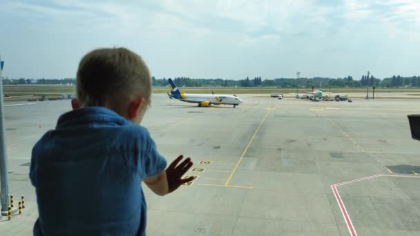 KIEV, UKRAINE-JULI 20, 2021: kleine passagier wacht op instappen op luchthaven, kind kijkt vliegtuigen in de buurt van een groot raam tijdens familie reizen — Stockvideo