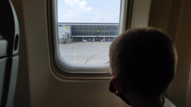 KIEV, UKRAINE-JÚLIUS 20, 2021: utazás világjárvány idején, maszkos gyermek utas néz ki a repülőgép ablakán vezetés közben a repülőtéren — Stock videók