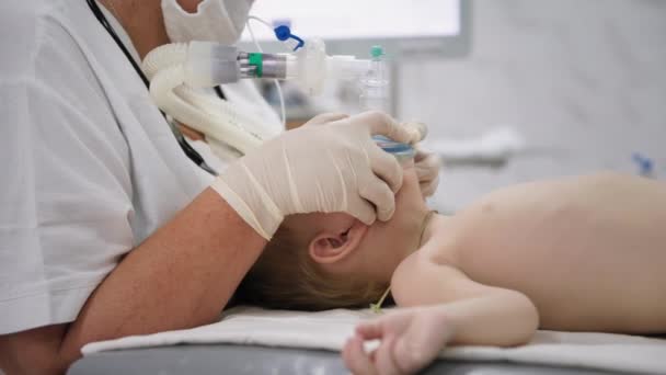 Child health, male kid nurse wearing gloves, holds an oxygen mask on his face to inject little patient into anesthesia before heart surgery in a hospital ward — Stock Video