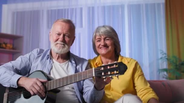 Gepensioneerd entertainment, mooi bejaard paar met een gitaar zwaaiende handen en praten in de voorkant van webcam thuis — Stockvideo