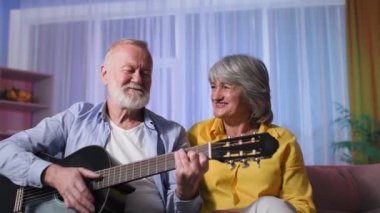 Yaşlı beyaz çift evde gitar çalarak eğleniyor. Emekli insanlar müzik aletleriyle eğleniyor.