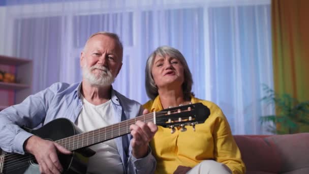 Ciesząc się życiem na emeryturze w domu, para seniorów grająca na gitarze akustycznej, starsza kobieta śpiewa ze swoim mężem gitarzystą siedzącym razem na kanapie — Wideo stockowe