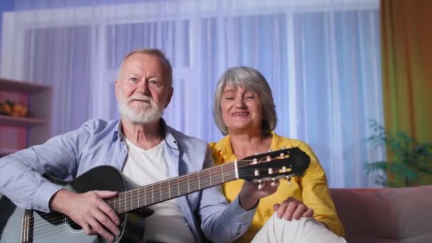 Siwe dziadkowie używać połączenia wideo za pośrednictwem laptopa kamery internetowej, kilka starych machając rękami i cieszyć się grając na gitarze siedzi na kanapie — Wideo stockowe