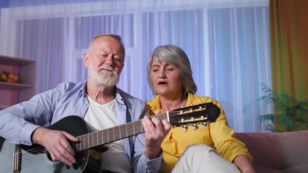 Vieux couple jouer de la guitare en ligne, mignon cheveux gris grands-parents chanter chanson sur appel vidéo via une webcam ordinateur portable tout en étant assis sur le canapé à la maison — Video