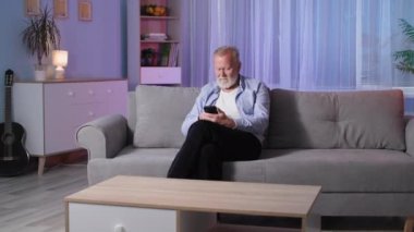 Evde aleti olan modern emekli, sakallı, yaşlı, kır saçlı adam koltukta oturan cep telefonunu kullanıyor, kamera hareket halinde.