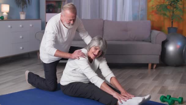 Mulher idosa fazendo exercício de alongamento com a ajuda do marido, casal de idosos fazendo esportes no tapete na sala de estar em casa — Vídeo de Stock