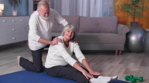 Aktiva pensionärer, sportig gamling träning träning hem, äldre make hjälper fru med full kropp stretching på mattan — Stockvideo