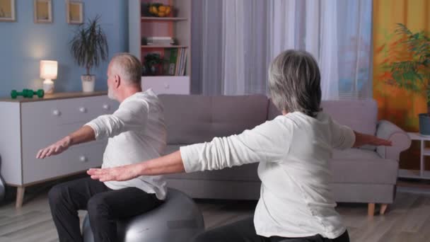Retirado aptitud, feliz sonriendo madura pareja de pelo gris juntos haciendo ejercicio sentado en fitballs en la habitación — Vídeo de stock