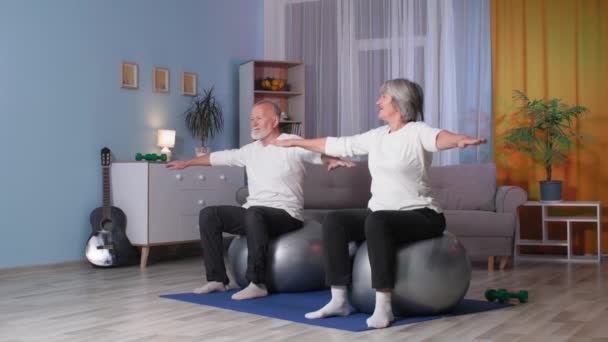 Gepensioneerden doen fitness op fitballen thuis, oudere grootouders met opgeheven armen draaien hun hele lichaam naar links en rechts — Stockvideo