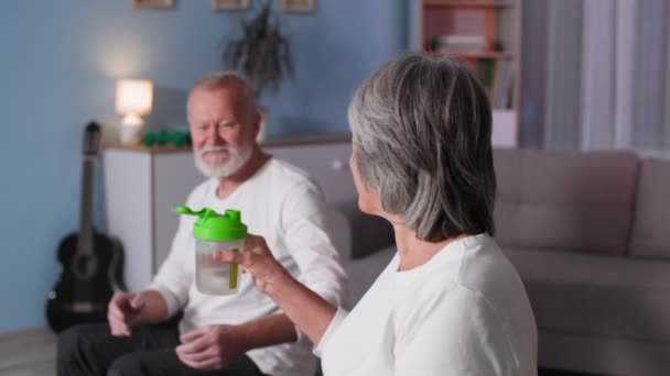 Спортивная пожилая пара, женщина пьет воду из бутылки после активных упражнений фитнес-тренировки с мужем в возрасте дома в комнате — стоковое видео