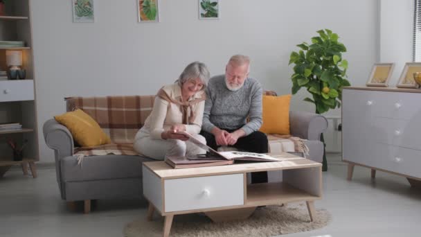 Recuerdos familiares, feliz pareja casada anciano hombre y mujer disfrutar de recuerdos volteando a través del viejo álbum familiar con fotos sentadas en el sofá en casa — Vídeo de stock