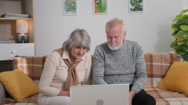 Pareja de ancianos consulta en línea, los abuelos de pelo gris están hablando en videollamada con el médico a través de una webcam portátil mientras está sentado en el sofá en casa — Vídeos de Stock