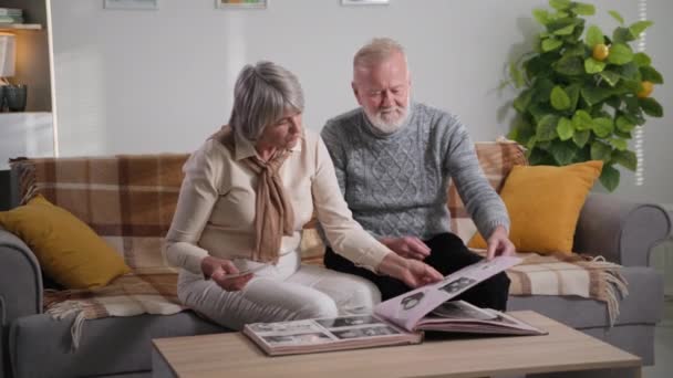 Elderly couple looks through old photographs, nostalgia and memories of youth and childhood, aged family looking album at home — Stockvideo