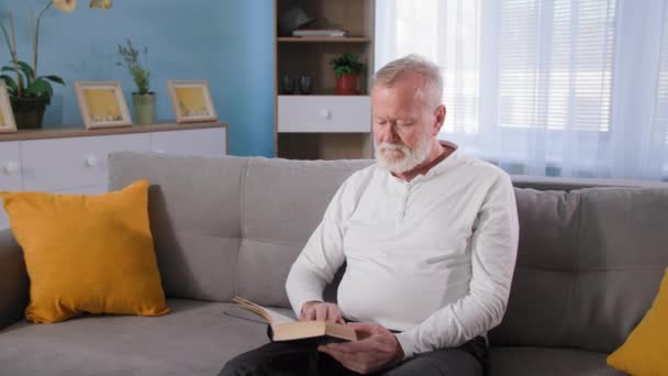 Livre de lecture de grand-père masculin âgé assis sur le canapé dans le salon, problème de santé chronique ou lombalgie chez les personnes âgées à la retraite — Video