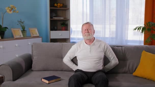 Emeryt cierpi na dyskomfort w dolnej części pleców, starszy, siwy mężczyzna z bólem pleców siedzi na kanapie w domu — Wideo stockowe
