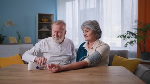 Äldre vårdande man kontrollerar blodtrycket på sin gamla fru med högt blodtryck med hjälp av en tonometer vid bordet, gamla par ta hand om hälsan hemma — Stockvideo