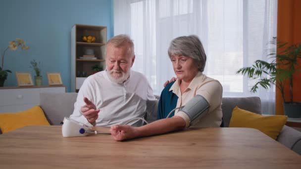 Boldog nyugdíjasok gondoskodnak az egészségükről, szerető férj méri a vérnyomást egy tonométerrel az idős feleségnek otthon — Stock videók