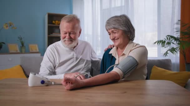 Porträtt av glada pensionärer som tar hand om hälsan, vårda äldre man mäta blodtryck till hustru med högt blodtryck i hemmet — Stockvideo