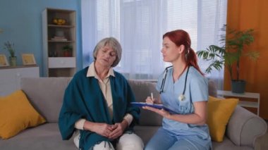 Profesyonel doktor ev ziyareti sırasında üst düzey kadın hastaya danışmanlık yapıyor ve not defterine not alıyor. Üniformalı mutlu bir terapist portresi ve kanepedeki gri saçlı kadın.