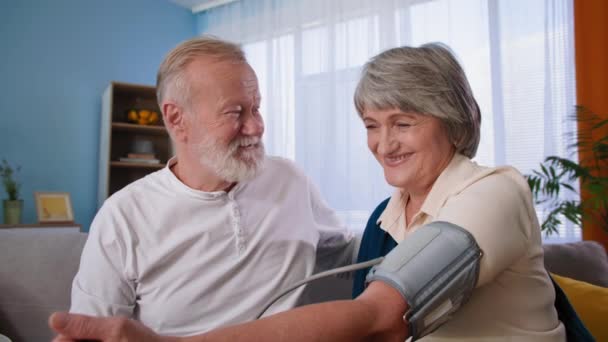 Gesundheitscheck zu Hause, reifer Mann kontrolliert Blutdruck seiner Frau mit Tonometer, Porträt glückliches älteres Paar — Stockvideo